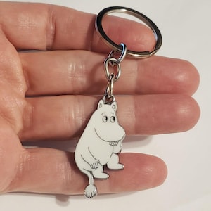 May include: A white enamel keychain featuring a cartoon character with a round body, small ears, and a tail. The character is outlined in black and attached to a silver keyring.