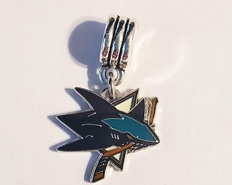 Pode incluir: Um berloque de metal prateado com um logótipo de tubarão em preto, azul-petróleo e branco, com um taco de hóquei castanho cruzado atrás do tubarão. O berloque inclui uma argola cilíndrica prateada no topo para prender a uma corrente.