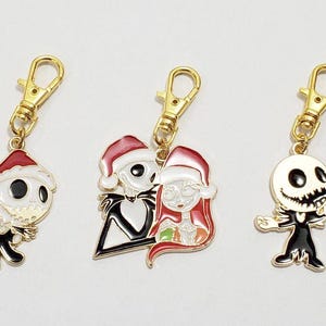 May include: Three gold-toned keychains featuring characters from The Nightmare Before Christmas. Each charm depicts Jack Skellington and Sally in Santa hats. The charms are black, white, red, and gold.