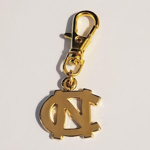 May include: A gold-colored charm with a lobster clasp. The charm features the interlocking "NC" logo, a symbol of the University of North Carolina. The charm is made of shiny metal and is designed to be attached to a keychain or bag.