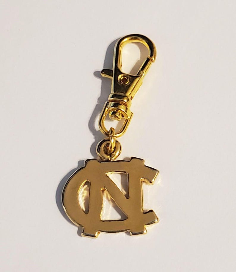 Op de afbeelding: Een goudkleurige bedel met een karabijnhaak. De bedel toont het in elkaar grijpende "NC"-logo, een symbool van de Universiteit van North Carolina. De bedel is gemaakt van glanzend metaal en is ontworpen om aan een sleutelhanger of tas te worden bevestigd.
