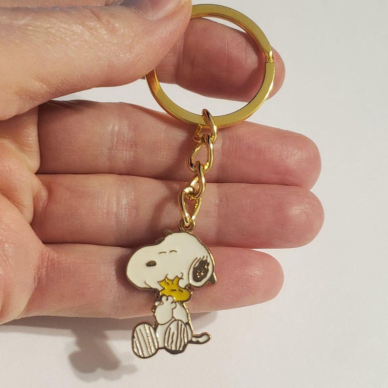 May include: A gold-colored keychain with a Snoopy charm. The charm features a white Snoopy with black spots, holding a yellow bird. The keychain has a gold ring and chain.