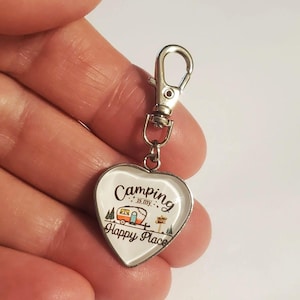 May include: Heart-shaped keychain with a silver clasp. The white heart features the text "Camping is my Happy Place" with an illustration of a camper, trees, and a signpost. The keychain is designed for camping enthusiasts.