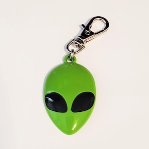May include: A bright green alien head keychain featuring a smooth, glossy finish and two large, solid black oval eyes. The pendant is attached to a silver-toned metal lobster claw clasp, providing a secure way to attach the item to keys or bags.