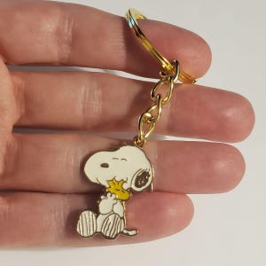 May include: A gold-colored keychain featuring a cartoon character. The character is white with black accents, holding a yellow bird. The keychain has a gold ring and chain.