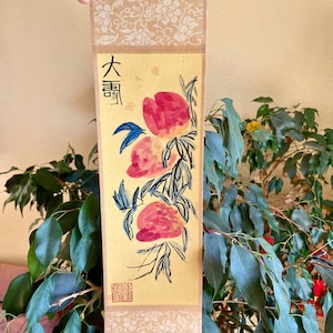 May include: A traditional Chinese scroll painting depicting three pink peaches on a branch with blue leaves. The scroll is mounted on a beige background with a floral pattern and includes the Chinese characters "大壽" (meaning "long life") and a red seal.