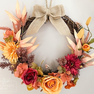 Könnte beinhalten: Ein saisonaler Kranz mit einem gewebten Weinreben-Sockel, verziert mit künstlichen Blumen in Orange-, Gelb- und Burgundertönen. Oben befindet sich eine Schleife aus Jute. Ideal für die Herbstdekoration.