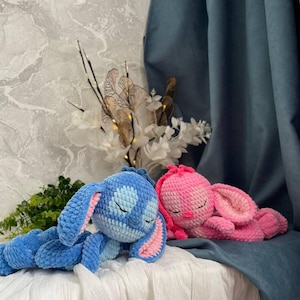 May include: Two handmade crochet bunny plushies. One is blue with pink inner ears, the other is pink with pink inner ears. Both are lying down with closed eyes, resting on a white cloth. The background includes a blue curtain and decorative flowers.