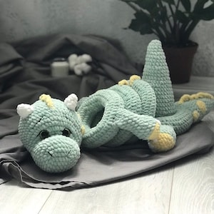 Può includere: Un giocattolo di drago verde all'uncinetto con accenti gialli e occhi bianchi. Il drago è rannicchiato su una coperta grigia.