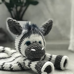 Puede incluir: Una manta de seguridad de peluche con forma de cebra hecha de hilo grueso y suave con rayas alternas blancas y gris oscuro. La cabeza de la cebra tiene un hocico gris oscuro, orejas puntiagudas, un mechón de pelo y ojos azules.