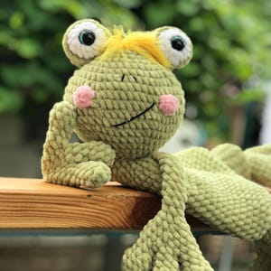 May include: A handmade, light green crochet frog plush toy with large, expressive eyes, pink cheeks, and a yellow tuft of hair. The frog is smiling and has long, floppy limbs. The plush is resting on a wooden surface.
