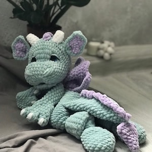 May include: A crocheted plush dragon toy with a light green body, purple wings, and white horns. The dragon is lying on a grey fabric surface.