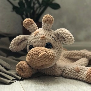 Könnte beinhalten: Ein handgefertigtes Giraffen-Plüschtier in Beige- und Brauntönen. Die weiche, gehäkelte Giraffe hat kleine, dunkelblaue Augen und detaillierte Merkmale, einschließlich Ohren und Hörner. Das Spielzeug liegt auf einer neutralfarbenen Oberfläche.