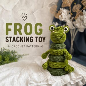 May include: A handmade crochet frog stacking toy in shades of green. The toy has a smiling face, arms, and legs, with stacking rings in varying shades of green. The text "FROG STACKING TOY CROCHET PATTERN" is displayed above the toy.