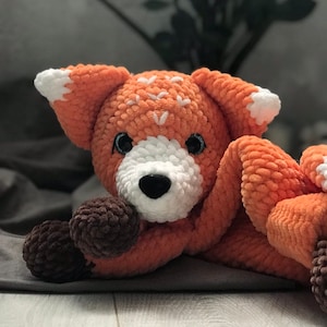 Puede incluir: Un peluche de zorro hecho a mano de ganchillo naranja y blanco. El zorro tiene un hocico, orejas y detalles blancos, con patas marrones oscuras y una nariz negra. El peluche está tumbado sobre una superficie gris.