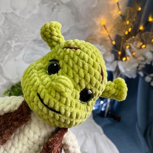 May include: A handmade, green crocheted doll resembling a cartoon ogre. The doll has large black eyes, a stitched smile, and small ears. It wears a cream-colored vest with brown accents. The doll is a whimsical, handcrafted item.
