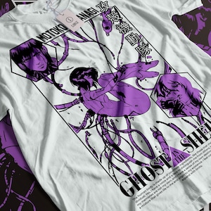 Może przedstawiać: Biały t-shirt z fioletowym i czarnym nadrukiem graficznym przedstawiającym kobietę z kablami i tekstem "MOTOKO ANAGI" i "GHOST IN THE SHELL".