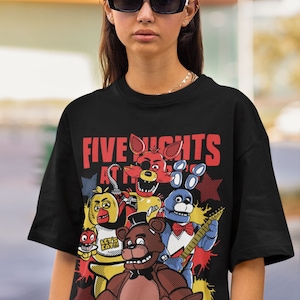Puede incluir: Camiseta negra con un estampado gráfico de personajes del videojuego Five Nights at Freddy's. El gráfico presenta a Freddy Fazbear, Bonnie the Bunny, Chica the Chicken, Foxy the Pirate Fox y un personaje más pequeño con un sombrero rojo. El texto "Five Nights at Freddy's" está impreso en rojo y blanco. El texto "Freddy Fazbear's Pizza" está impreso en blanco con un contorno negro. El texto "Where fantasy and fun come to life" está impreso en blanco debajo del logotipo de la pizzería.
