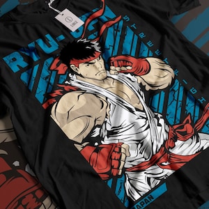 Puede incluir: Camiseta negra con un gráfico de Ryu de Street Fighter. El diseño incluye el texto "RYU-SAN" y "STREET FIGHTER" en azul, con una diadema y guantes rojos. La etiqueta indica una talla "L".