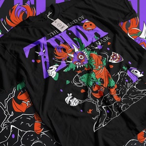 Puede incluir: Camiseta negra con un gráfico morado y blanco de Majora's Mask de The Legend of Zelda: Majora's Mask. El gráfico presenta la máscara con una expresión amenazante y un atuendo verde y naranja.