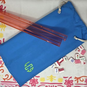 May include: A blue drawstring bag with a green monogram, a clear orange tile rack, and several mahjong tiles. The tiles are white with red, pink, and green designs. The bag is on a patterned white and pink surface.