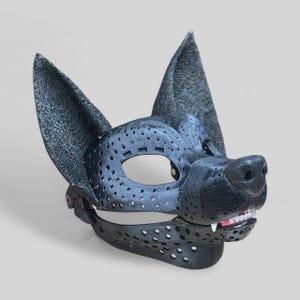 Puede incluir: Una máscara de perro negra con orejas grandes y erguidas y un hocico detallado. La máscara tiene numerosos agujeros pequeños y una mandíbula inferior articulada que revela dientes blancos y encías rosas. Las orejas tienen una tela gris texturizada.