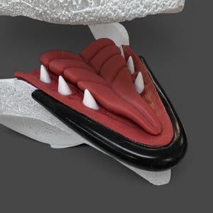 May include: A 3D printed creature jaw model featuring a white textured upper structure, a dark red tongue with segmented detailing, sharp white conical teeth, and a smooth black lower jaw rim, presented against a solid dark grey background.