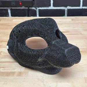 May include: A black, textured animal mask with large eye openings and a snout. The mask is made of a woven material and appears to be 3D-printed. The mask is designed to fit over the head and face.