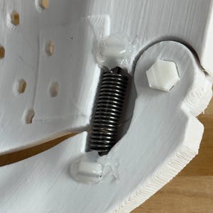 May include: A white 3D-printed component featuring a textured surface with a series of small hexagonal holes. A metallic silver coil spring is secured between two sections using clear adhesive, alongside a white hexagonal bolt fastener.