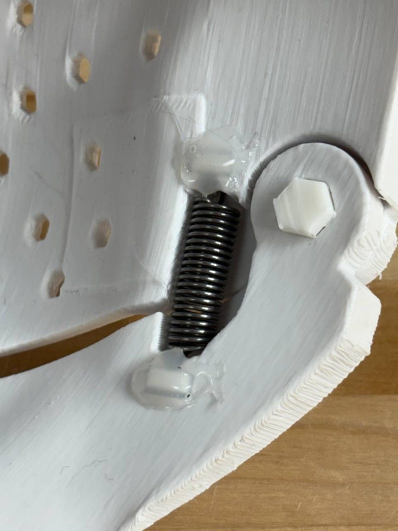 May include: A white 3D-printed component featuring a textured surface with a series of small hexagonal holes. A metallic silver coil spring is secured between two sections using clear adhesive, alongside a white hexagonal bolt fastener.