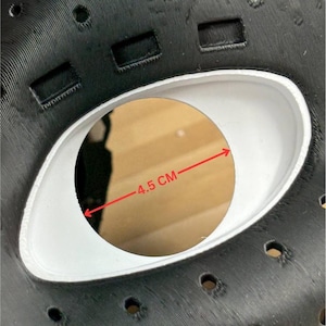 May include: A black mask with a textured surface featuring multiple small circular ventilation holes and rectangular slots. A central white eye opening is visible, with a red arrow indicating a measurement of 4.5 cm across the opening.