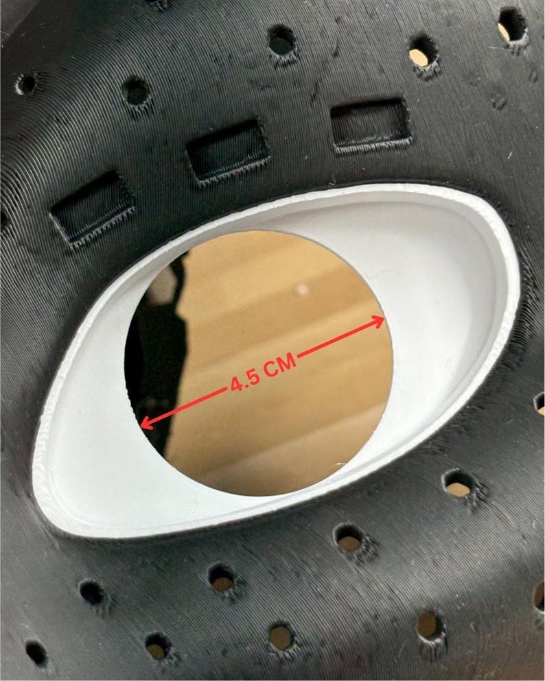 May include: A black mask with a textured surface featuring multiple small circular ventilation holes and rectangular slots. A central white eye opening is visible, with a red arrow indicating a measurement of 4.5 cm across the opening.