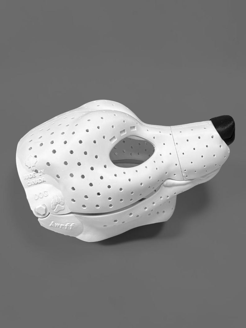 May include: A white, perforated dog-shaped mask with a black nose. The side features embossed text reading "MADE IN CANADA", "DOG", and "Awrff", along with a paw print icon. The mask has a hollow eye opening and a smooth, molded structure.