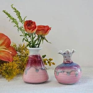 May include: Two ceramic vases, one conical and one round, in shades of pink, red, and blue. The vases hold orange roses and greenery. The background is a soft white, creating a clean and inviting aesthetic.