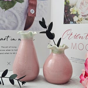 May include: Two pink ceramic vases with ruffled rims and a light green trim. The vases hold black decorative stems. The background includes a floral print and text that reads "Oriental MOOD."