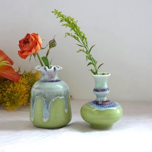 May include: Two ceramic vases in shades of green, with a ruffled rim on one. The vases hold a rose and greenery. The vases have a glossy finish with blue and purple accents. The background is a soft white.