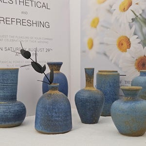May include: A collection of blue ceramic vases with a textured finish and a contrasting tan rim. The vases vary in shape and size, with some featuring a rounded body and others a cylindrical design. The background includes a wedding invitation and floral imagery.