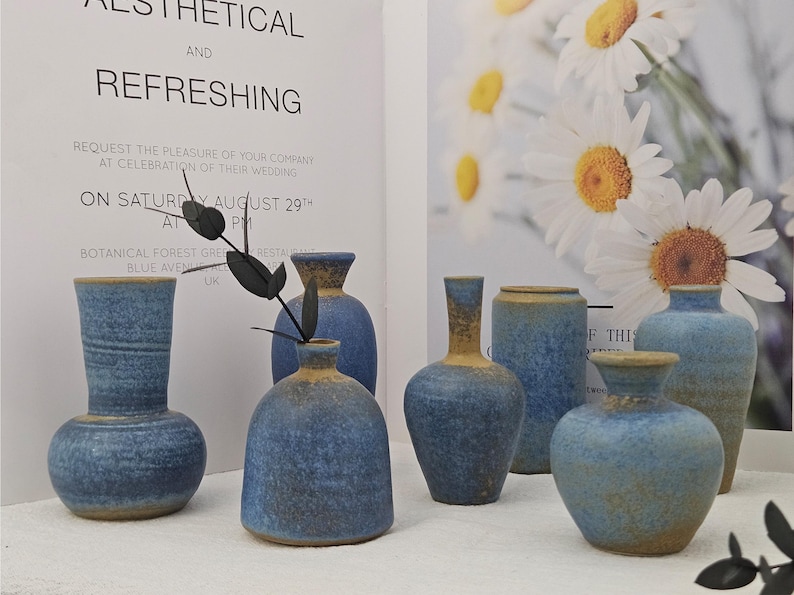 May include: A collection of blue ceramic vases with a textured finish and a contrasting tan rim. The vases vary in shape and size, with some featuring a rounded body and others a cylindrical design. The background includes a wedding invitation and floral imagery.