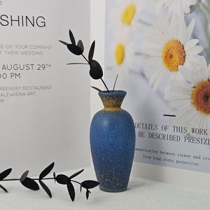 May include: A blue ceramic vase with a yellow rim holds a black decorative plant. The vase is set against a backdrop of a wedding invitation and a floral print. The invitation reads "Saturday August 29th at 4:00 PM".