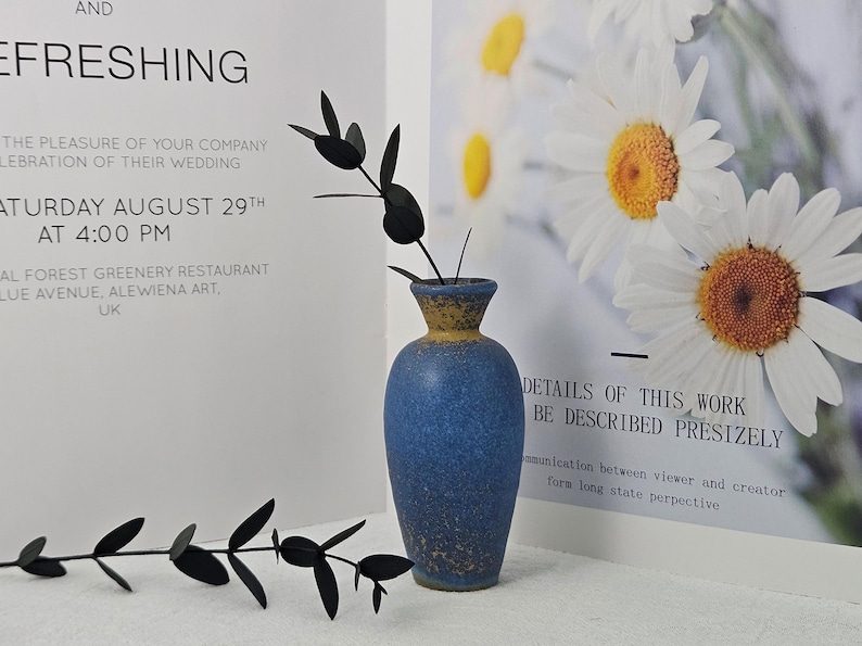May include: A blue ceramic vase with a yellow rim holds a black decorative plant. The vase is set against a backdrop of a wedding invitation and a floral print. The invitation reads "Saturday August 29th at 4:00 PM".