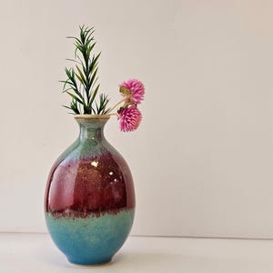 Może przedstawiać: Ceramiczny wazon z niebieską, zieloną i czerwoną glazurą. Wazon jest okrągły i ma wąską szyjkę. Jest wypełniony gałązką zielonych liści i różowymi kwiatami.