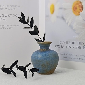 May include: A blue ceramic vase with a textured surface, holding black leaf stems. The vase is set against a white background with printed text and a floral image. The text reads "DETAILS OF THIS WORK CAN BE DESCRIBED PRESIZELY."