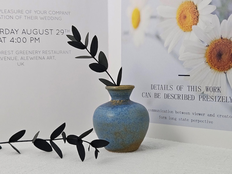 May include: A blue ceramic vase with a textured surface, holding black leaf stems. The vase is set against a white background with printed text and a floral image. The text reads "DETAILS OF THIS WORK CAN BE DESCRIBED PRESIZELY."