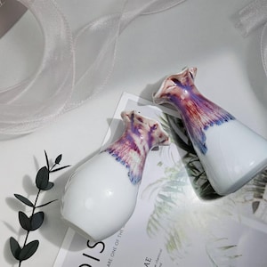 May include: Two white ceramic vases with a purple and pink floral design. The vases are on a white surface with a white fabric in the background.