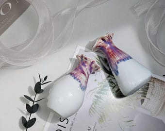 Puede incluir: Dos jarrones de cerámica blanca con un diseño floral morado y rosa. Los jarrones están sobre una superficie blanca con una tela blanca en el fondo.
