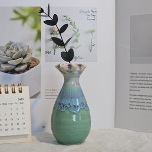 K&ouml;nnte beinhalten: Eine Keramikvase mit gr&uuml;n-blauer Glasur, die einen schwarzen dekorativen Zweig h&auml;lt. Ein Kalender mit Mai 2025 ist im Vordergrund. Die Vase steht auf einer wei&szlig;en Oberfl&auml;che, mit einem offenen Buch im Hintergrund.