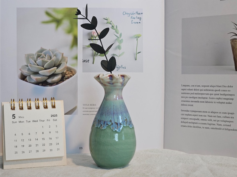 K&ouml;nnte beinhalten: Eine Keramikvase mit gr&uuml;n-blauer Glasur, die einen schwarzen dekorativen Zweig h&auml;lt. Ein Kalender mit Mai 2025 ist im Vordergrund. Die Vase steht auf einer wei&szlig;en Oberfl&auml;che, mit einem offenen Buch im Hintergrund.
