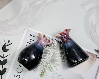 May include: Two ceramic vases with a glossy finish. The vases have a dark blue base that fades to a lighter blue and then to a purple and cream-colored top. The vases are on a white surface with a magazine and decorative elements.