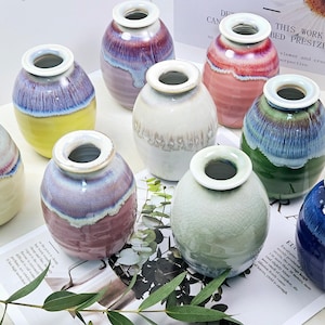 May include: A collection of ten small ceramic vases in various colours and patterns. The vases are all round and have a smooth, glossy finish. They are arranged in a group on a white surface.