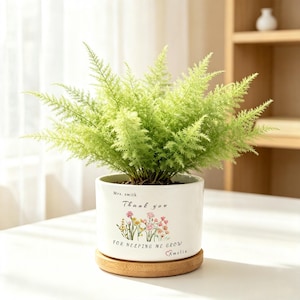 May include: A white ceramic cylindrical planter containing a lush, light green asparagus fern. The planter features the printed text "Mrs. smith Thank you FOR HELPING ME GROW Amelia" with a small red heart and a colourful floral illustration.
